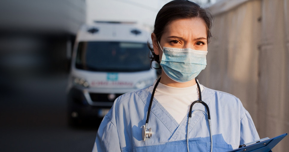A nurse wearing a mask and a stethoscope around her neck is looking into the camera.