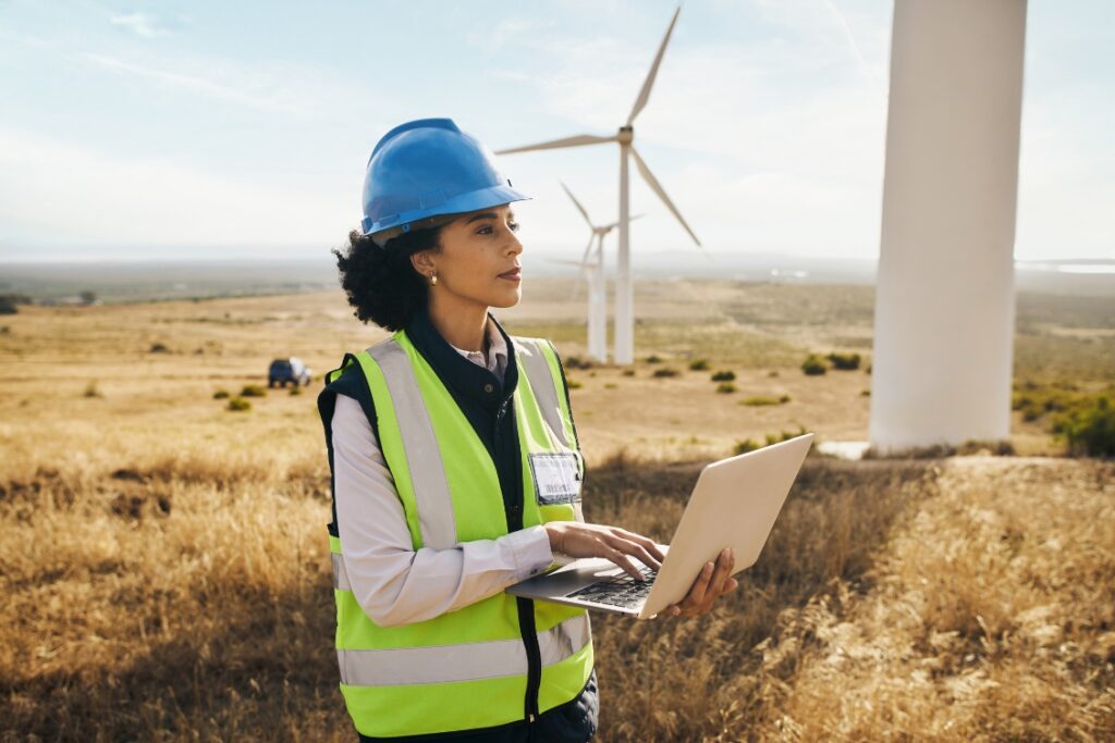 A woman in a vest and hard hat holding a computer in a field of wind turbines.